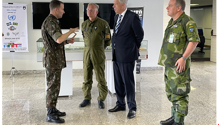 Quatro homens em pé. Três deles à direita escutam um à esquerda. Na ordem, da esquerda para a direita, dois militares, um civil e outro militar.