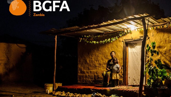 Young girl standing next to her mum outside their house. It is dark outside and a solar powered light bulb is lighting up their veranda.